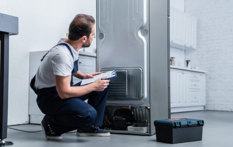 handyman in working overall writing in clipboard near broken refrigerator in kitchen